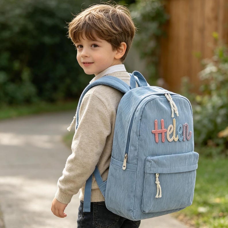Puede incluir: Una mochila de pana azul claro con el nombre "Helen" bordado en letras coloridas. La mochila tiene un bolsillo frontal, un asa superior y correas ajustables. El ni&ntilde;o lleva un su&eacute;ter beige y vaqueros oscuros.