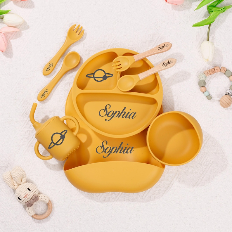 May include: Mustard yellow baby feeding set with the name "Sophia" printed on the plate and bib. The set includes a divided plate, bowl, cup, bib, and utensils. A planet graphic is on the plate and cup.