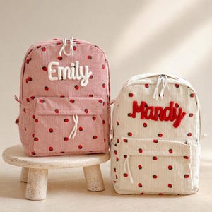 Puede incluir: Dos mochilas de pana, una rosa y otra crema, adornadas con patrones de fresas rojas. La mochila rosa presenta el nombre "Emily" en blanco, mientras que la crema muestra "Mandy" en rojo. Ambas tienen bolsillos delanteros y cremalleras blancas.