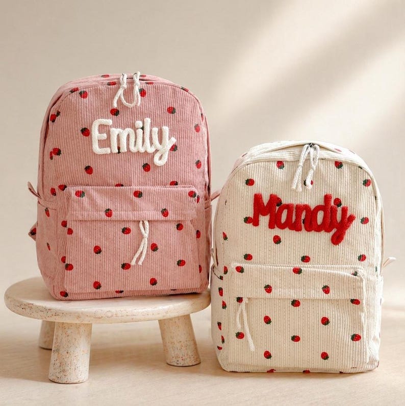 Puede incluir: Dos mochilas de pana, una rosa y otra crema, adornadas con patrones de fresas rojas. La mochila rosa presenta el nombre "Emily" en blanco, mientras que la crema muestra "Mandy" en rojo. Ambas tienen bolsillos delanteros y cremalleras blancas.