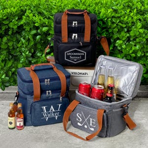 Peut inclure: Trois sacs isothermes noirs, bleu marine et gris, avec des poignées et des sangles marron. Le sac noir porte l'inscription "GROOMSMAN Samuel". Le sac bleu marine porte l'inscription "TAT Walker". Le sac gris porte "SxE" avec des clubs de golf croisés.