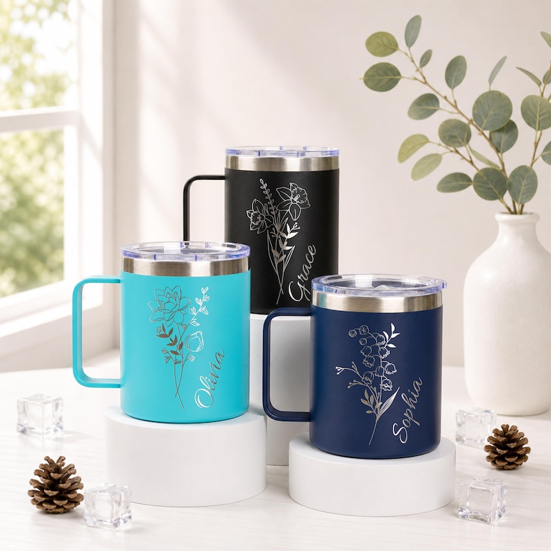 May include: Three insulated travel mugs in teal, black, and navy blue, each with a silver floral design and a name. The mugs have silver lids and handles. The teal mug says "Olivia". The black mug says "Grace". The navy mug says "Sophia".