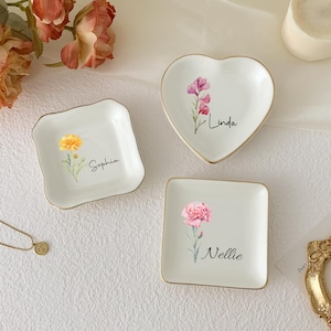 May include: Three white ceramic trinket dishes in square and heart shapes, each with a gold rim and a floral design. The dishes are personalized with names: "Sophia", "Linda", and "Nellie".