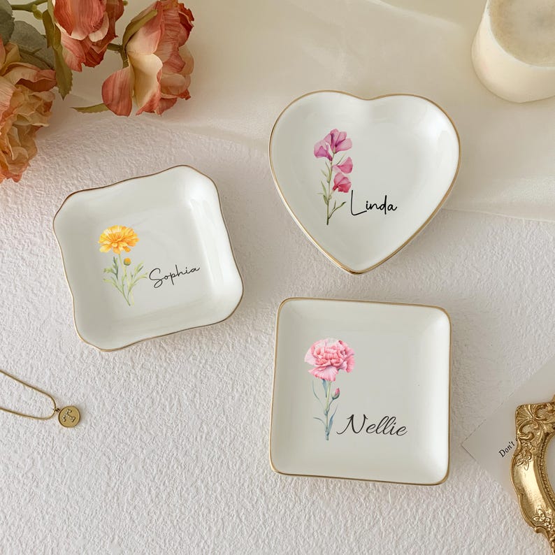 May include: Three white ceramic trinket dishes in square and heart shapes, each with a gold rim and a floral design. The dishes are personalized with names: "Sophia", "Linda", and "Nellie".