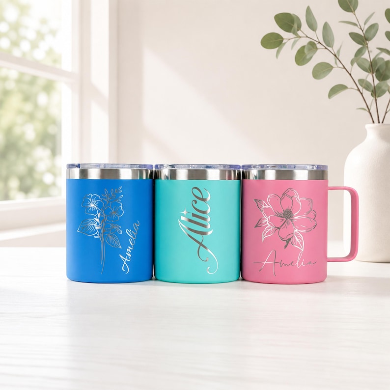May include: Three insulated travel mugs in blue, teal, and pink, each with a silver floral design and a name. The blue mug has a floral design and the name "Amelia". The teal mug has the name "Alice". The pink mug has a magnolia and the name "Amelia".