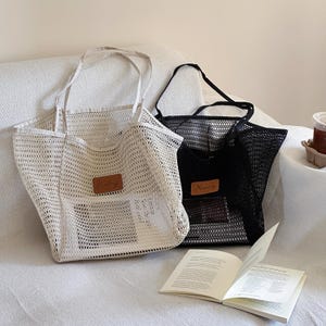 May include: Two mesh tote bags, one cream and one black, with long handles. Each bag has a rectangular leather patch with a name on it. A book is open in front of the bags.