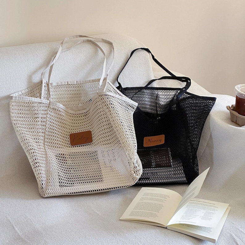 May include: Two mesh tote bags, one cream and one black, with long handles. Each bag has a rectangular leather patch with a name on it. A book is open in front of the bags.