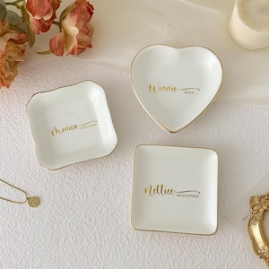 May include: Three white ceramic ring dishes with gold trim. One is heart-shaped with "Winnie Bride" in gold script. Another is square with "Nettice Bridesmaid". The third is a square shape with "Monica Bridesmaid".