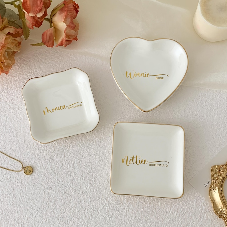May include: Three white ceramic ring dishes with gold trim. One is heart-shaped with "Winnie Bride" in gold script. Another is square with "Nettice Bridesmaid". The third is a square shape with "Monica Bridesmaid".