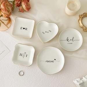 May include: A collection of white ceramic ring dishes in various shapes: square, heart, and round. Each dish has a gold rim and personalized text in black, including initials, names, and a date. A silver ring is also present.