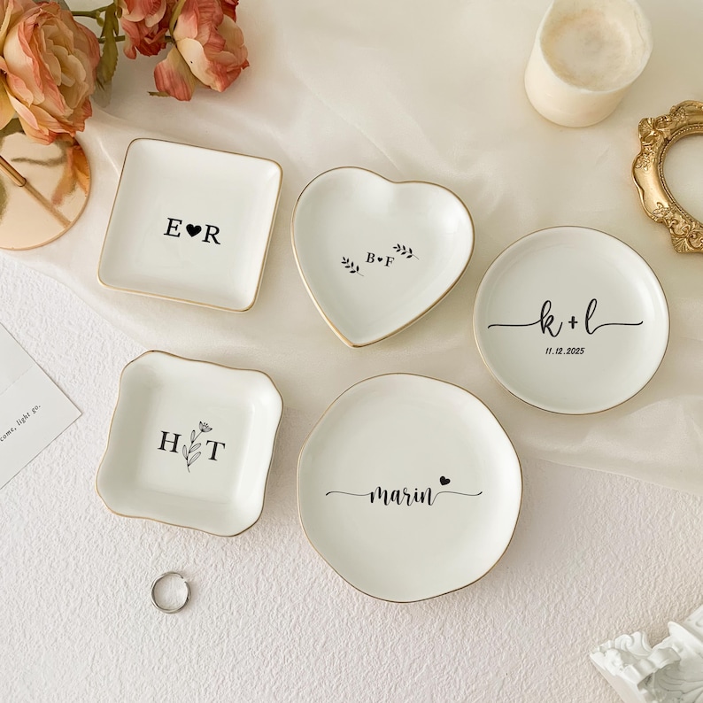 May include: A collection of white ceramic ring dishes in various shapes: square, heart, and round. Each dish has a gold rim and personalized text in black, including initials, names, and a date. A silver ring is also present.