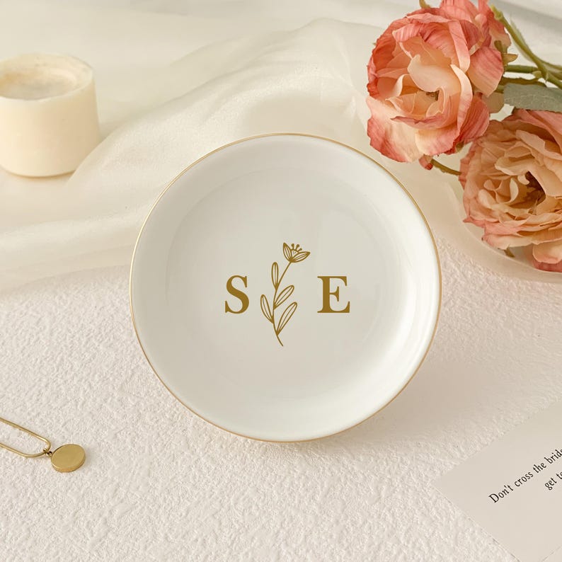 May include: A white ceramic ring dish with a gold rim. The dish features the gold initials "S E" with a floral design. A gold necklace and a candle are in the background. Pink flowers are also visible.