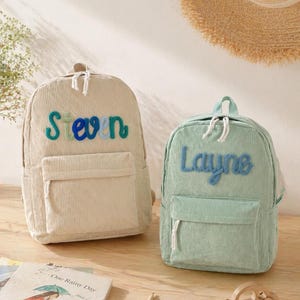Puede incluir: Dos mochilas de pana, una beige y otra verde menta, se muestran juntas. La mochila beige tiene el nombre "Steven" bordado en verde azulado y azul. La mochila verde menta tiene el nombre "Layne" bordado en azul. Ambas tienen un bolsillo frontal.