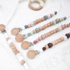 May include: Baby pacifier clips with wooden beads, silicone beads, and metal clips. The clips are personalised with names. The colour palette includes grey, pink, and brown. A wooden teething ring with a knitted cover is also visible.