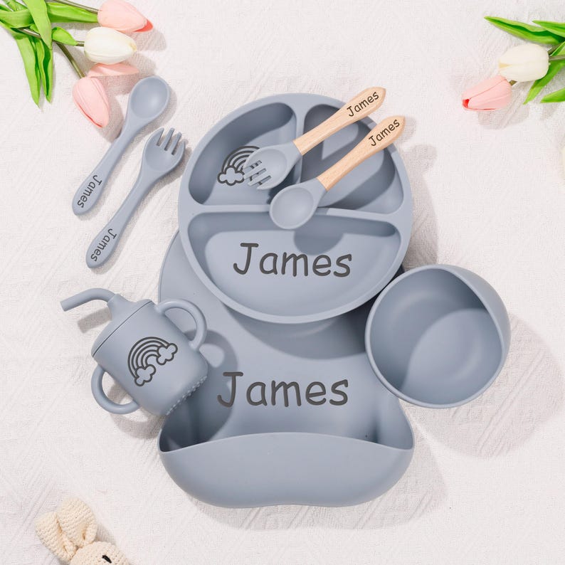 May include: A baby feeding set in a soft gray color, featuring a divided plate, bowl, bib, sippy cup, and utensils. The set includes a spoon, fork, and two wooden-handled spoons. Each item is personalized with the name "James" and a rainbow graphic.