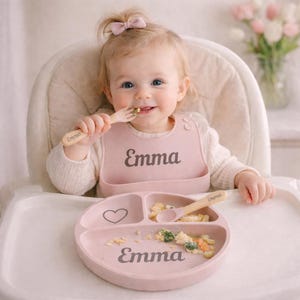 May include: A baby is seated in a high chair, wearing a pink bib and using a wooden fork and spoon. The bib, plate, and utensils are personalized with the name "Emma." The plate has three sections and a heart design.