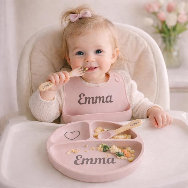 May include: A baby is seated in a high chair, wearing a pink bib and using a wooden fork and spoon. The bib, plate, and utensils are personalized with the name "Emma." The plate has three sections and a heart design.