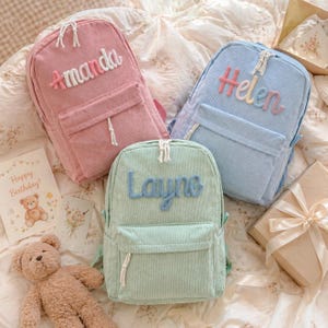 Puede incluir: Tres mochilas de pana en rosa, azul claro y verde menta, cada una con un nombre bordado en la parte delantera. La mochila rosa dice "Amanda", la azul dice "Helen" y la verde dice "Layne". Cada una tiene un bolsillo delantero y un cord&oacute;n blanco.
