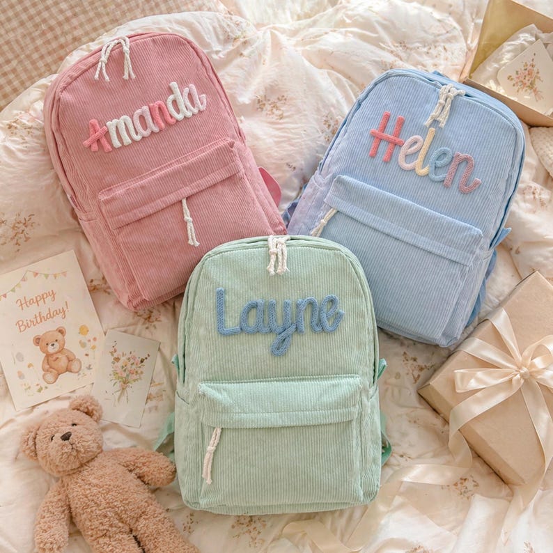 Puede incluir: Tres mochilas de pana en rosa, azul claro y verde menta, cada una con un nombre bordado en la parte delantera. La mochila rosa dice "Amanda", la azul dice "Helen" y la verde dice "Layne". Cada una tiene un bolsillo delantero y un cord&oacute;n blanco.