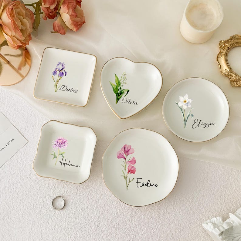 May include: A collection of small, white ceramic dishes in various shapes: square, heart, and round. Each dish features a floral design and a name in script. The edges are trimmed with gold. The flowers include irises, lilies of the valley, and sweet peas.
