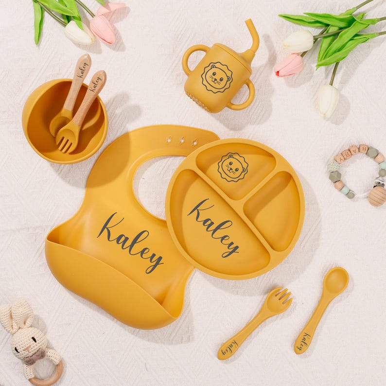 May include: Mustard yellow baby feeding set featuring a divided plate, bib, bowl, cup with straw, spoon, and fork. The set includes a lion face design and the name "Kaley" in black script. Wooden utensils are included.