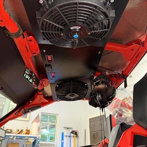 May include: Black panel with two electric fans, a digital display, and a USB port, mounted on a red frame. The fans are circular with black blades and a blue center. The panel is installed in a vehicle.