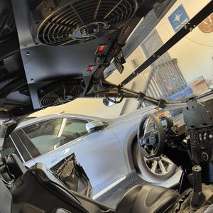 May include: Interior view of a black off-road vehicle with two large cooling fans mounted on the ceiling. The vehicle has black seats, a steering wheel, and a dashboard with various controls. A car is visible in the background.