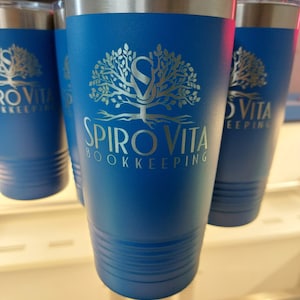 May include: Blue insulated tumblers with silver lids. The tumblers feature a gold-colored tree logo with the text "Spiro Vita Bookkeeping". The tumblers are cylindrical and appear to be made of stainless steel.