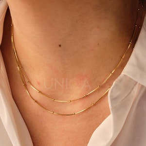 May include: A gold chain necklace with two strands of small, round beads and thin, straight bars. The necklace is worn by a person with a white shirt.