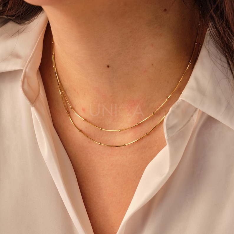 May include: A gold chain necklace with two strands of small, round beads and thin, straight bars. The necklace is worn by a person with a white shirt.