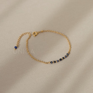 May include: A fine gold-coloured bracelet with small, dark blue beads. The bracelet has a delicate chain design and an adjustable clasp. The background is a soft, neutral colour.