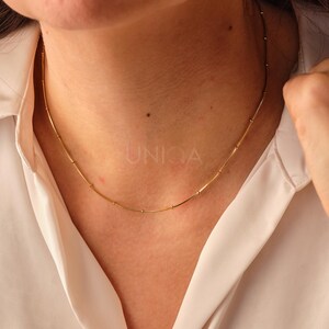 May include: A gold chain necklace with small round beads and longer, thin bars connecting them. The necklace is worn by a person with a white shirt.
