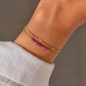 Puede incluir: Un brazalete de cadena dorada con una cadena delicada y una fila de pequeñas cuentas rosas.