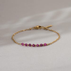 Könnte beinhalten: Ein zartes, goldfarbenes Armband mit einer feinen Kette und einem einfachen Verschluss. Es zeigt eine Reihe kleiner, runder, rubinroter Perlen. Die Perlen sind gleichmäßig entlang der Kette angeordnet und schaffen ein minimalistisches und elegantes Design.