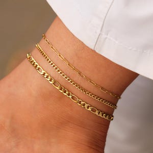Könnte beinhalten: Drei goldene Ketten-Fußkettchen, die an einem Knöchel übereinander liegen. Die Fußkettchen haben unterschiedliche Breiten und Stile.