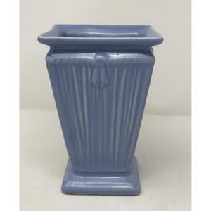 Può includere: Un vaso in ceramica blu pervinca di forma quadrata con bordo svasato e base. L'esterno presenta dettagli a coste verticali e una fascia decorativa simile a una corda che circonda la sezione superiore, creando un aspetto strutturato e uniforme.