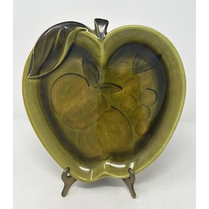 以下が含まれることがあります： 茶色い茎と葉が付いた、緑色のセラミック製リンゴ型サービング皿。茶色と緑の色合いで描かれたフルーツのデザインが特徴です。ブロンズ色の金属製スタンドに飾られています。