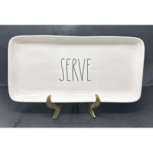 May include: A rectangular white ceramic serving platter with rounded corners. The word "SERVE" is printed in black, centered on the platter. The platter is displayed on a decorative gold-colored metal stand.