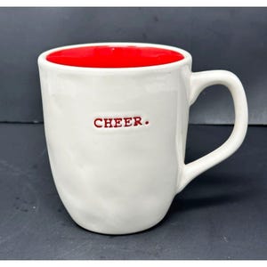Puede incluir: Taza de cerámica blanca con interior rojo y asa curva. La palabra "CHEER." está impresa en rojo en la parte delantera de la taza. La taza tiene una superficie ligeramente texturizada.