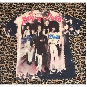 Puede incluir: Una camiseta de manga corta con un efecto de desteñido con lejía en blanco y negro que presenta un gráfico de la banda New York Dolls. El texto New York Dolls aparece en rosa en la parte superior y en azul en el centro de la imagen.