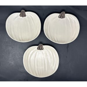 May include: Three off-white ceramic plates shaped like pumpkins. Each plate features a glossy finish, vertical ridges mimicking the texture of a pumpkin, and a small, dark gray stem detail at the top center of the rim, arranged in a triangular pattern.