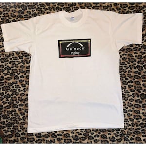 Peut inclure: Un t-shirt blanc à col rond et manches courtes. Le t-shirt présente un graphisme rectangulaire sur le devant avec le texte "AIRTOUCH" au-dessus de "Paging" dans un encadré noir avec des bordures rouges, jaunes et blanches. Le t-shirt est en matière souple.