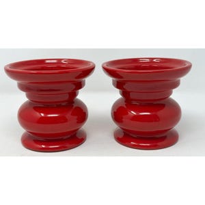 May include: A pair of matching red ceramic candle holders. Each has a wide, shallow top, a rounded middle section, and a flared base. The glossy finish reflects light, highlighting the smooth curves and vibrant colour. These decorative pieces are suitable for home decor.