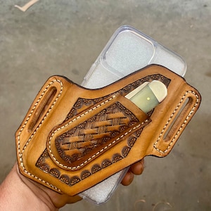 May include: A brown leather phone case with intricate tooled designs and a pocket for a small knife. The case is designed to fit a smartphone with a clear plastic back.
