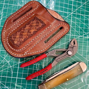 May include: A brown leather sheath with decorative stitching and tooling, holding a pair of red-handled pliers and a folding knife with a light-colored handle. The tools are on a green cutting mat.