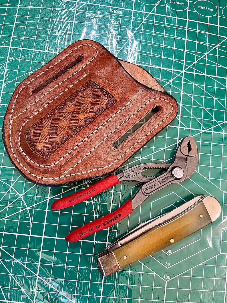 May include: A brown leather sheath with decorative stitching and tooling, holding a pair of red-handled pliers and a folding knife with a light-colored handle. The tools are on a green cutting mat.