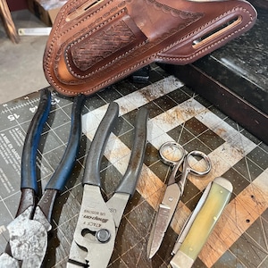 May include: A brown leather holster with decorative tooling, alongside various metal tools including pliers, scissors, and a folding knife. The tools are arranged on a textured surface.