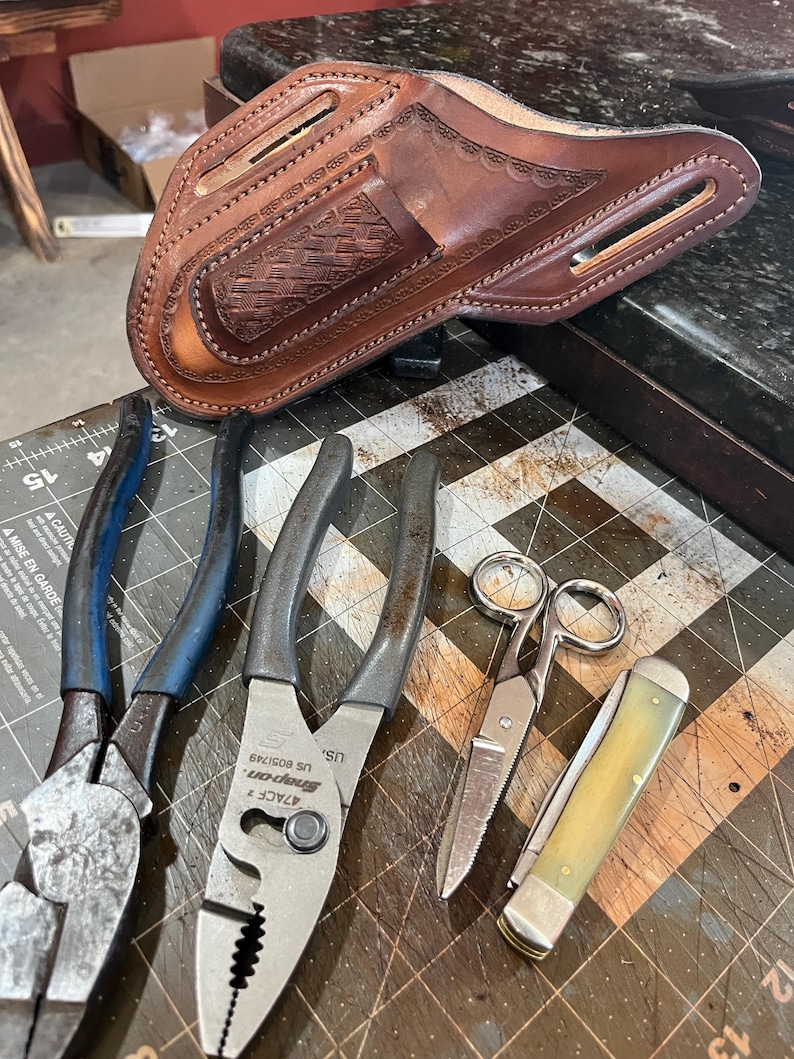 May include: A brown leather holster with decorative tooling, alongside various metal tools including pliers, scissors, and a folding knife. The tools are arranged on a textured surface.