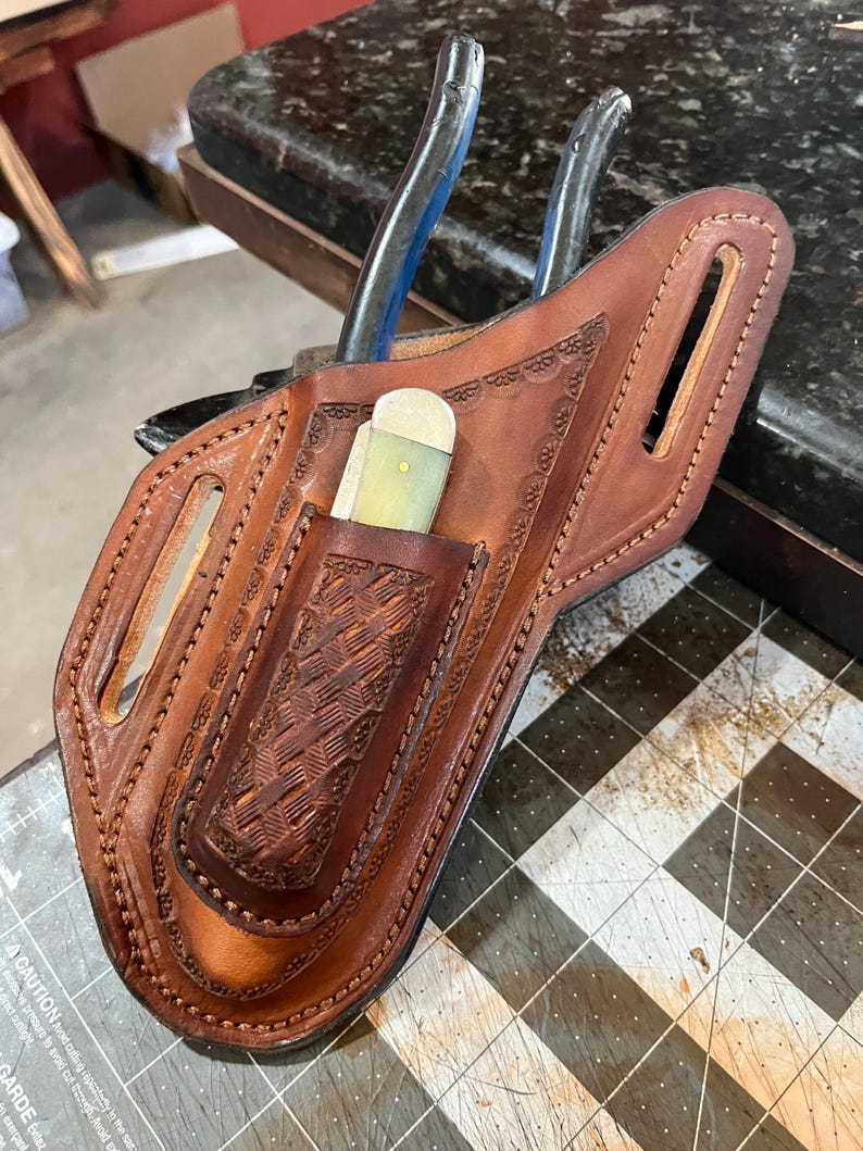May include: A brown leather tool holster with an embossed pattern. The holster has a pocket holding a small knife and slots for tools. A pair of pliers is partially inserted into the holster.