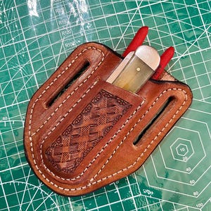 Puede incluir: Una funda de cuero marrón para cuchillo con un patrón decorativo en relieve y costuras blancas. Un cuchillo con mango de color claro y detalles rojos es parcialmente visible. La funda está sobre una esterilla de corte verde.